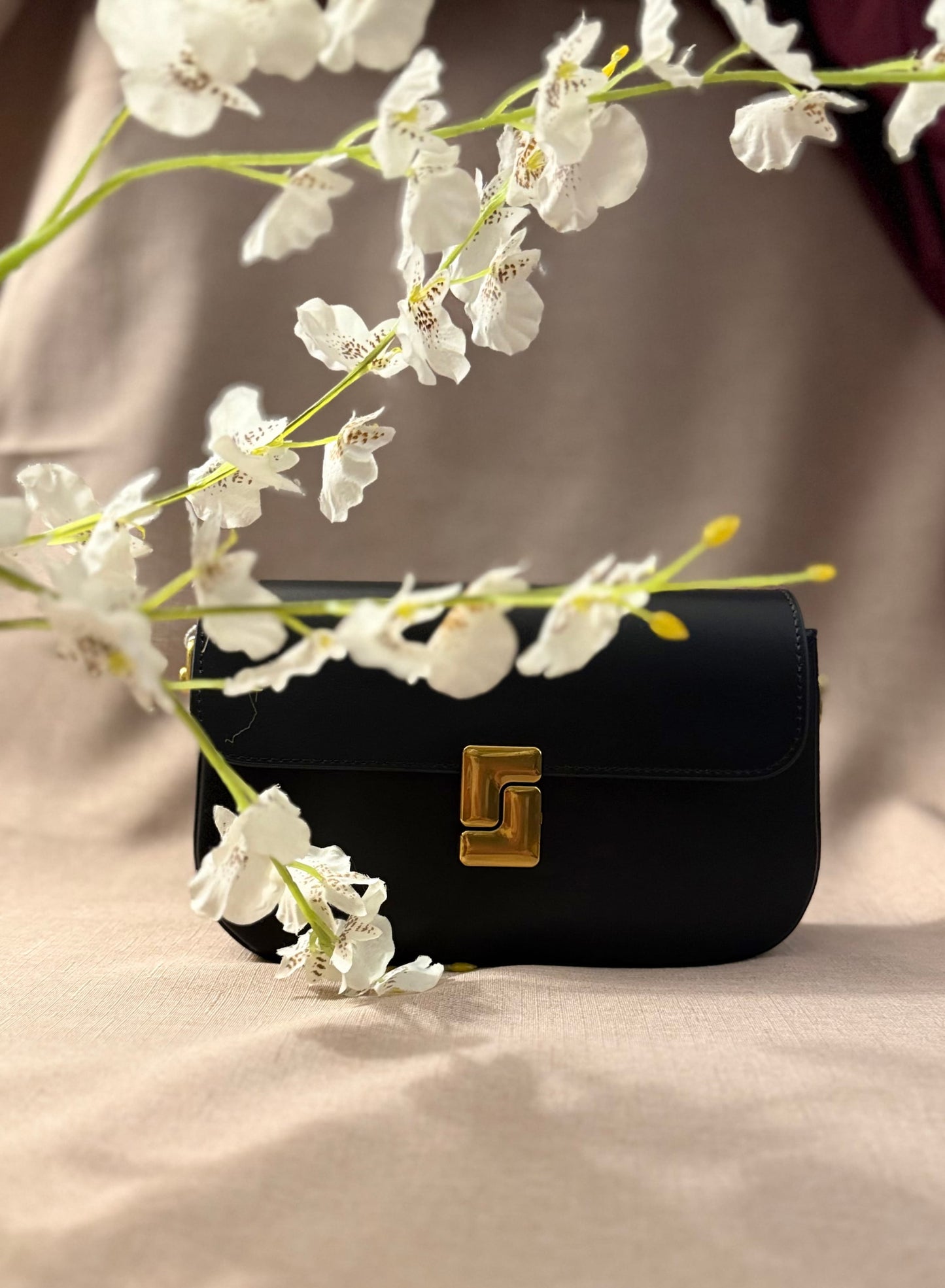 Black handbag with gold clasp on a beige surface with white flowers